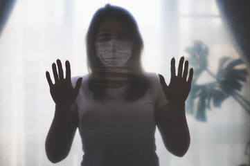 The silhouette of a girl in a medical mask, in quarantine, standing in a room with her hands leaning against a plastic film barrier.