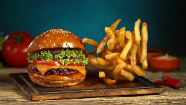 Super Slow Motion Shot of Fast Food Concept. Falling French Fries on Wooden Cutting Board next to the Fresh Hamburger at 1000fps.