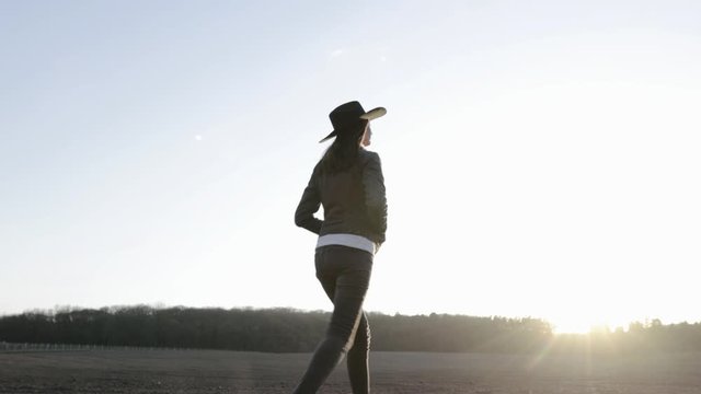 Woman walking lonely through an empty field at sunset wearing cawboy hat and jeans coat. She is smiling and enjoying spring.