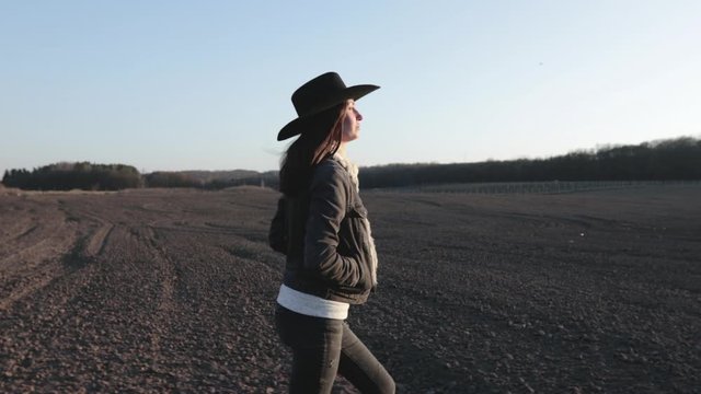 Woman walking lonely through an empty field at sunset wearing cawboy hat and jeans coat. She is smiling and enjoying spring.