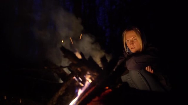 Woman traveler sitting near a fire and looking at her travel photo on a smartphone at night. A tourist girl enjoys a warm, cozy summer evening near a bonfire in nature.