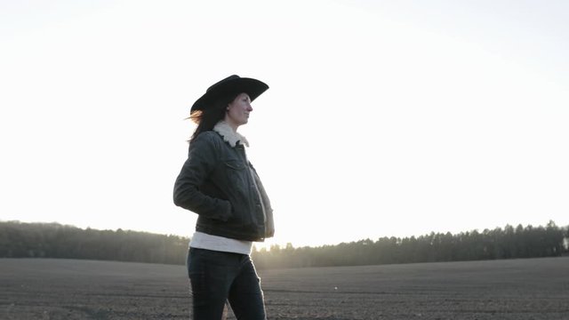 Woman walking lonely through an empty field at sunset wearing cawboy hat and jeans coat. She is smiling and enjoying spring.