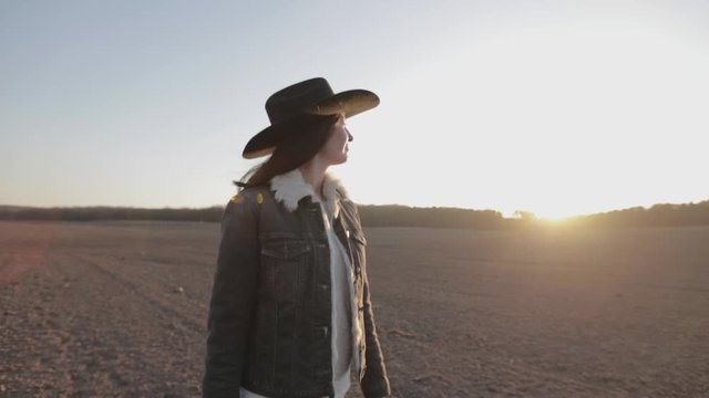 Woman walking lonely through an empty field at sunset wearing cawboy hat and jeans coat. She is smiling and enjoying spring.