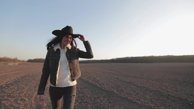 Woman walking lonely through an empty field at sunset wearing cawboy hat and jeans coat. She is smiling and enjoying spring.