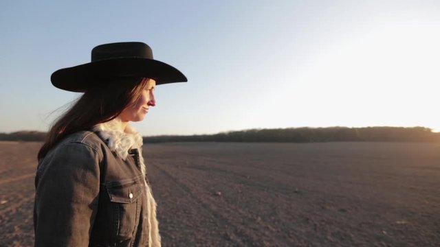 Woman walking lonely through an empty field at sunset wearing cawboy hat and jeans coat. She is smiling and enjoying spring.