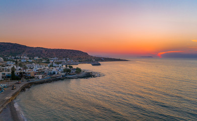Fototapeta premium Gorgeus sunset over sea with waves, rocks and traditional greek village of Milatos, Crete, Greece.