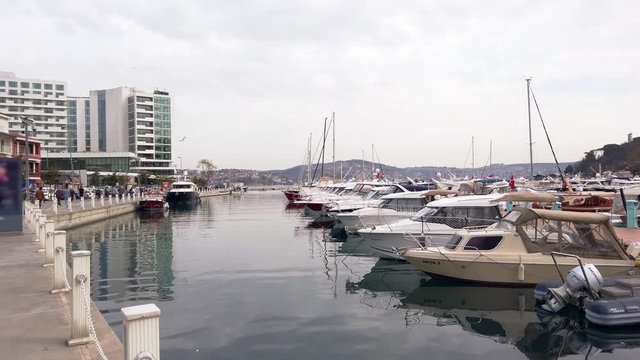 Cloudy foggy day. Private yachts and sailing boats with lowered sails standing on pier in bay. Reflections in water. People walk along promenade. Turkey, Istanbul, Tarabya, March 2020.