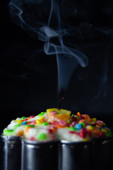 Birthday cupcake with colored sweets and candles in a cloud of smoke on a dark background