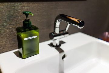 New modern faucet in bathroom closeup with stainless steel handle and white sink with green hand soap dispenser sign and dark background