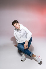 Pensive teen boy posing sitting on the floor in the studio. Portrait of stylishly dressed student. Studio shot Handsome attractive european man sitting on floor on a pink background