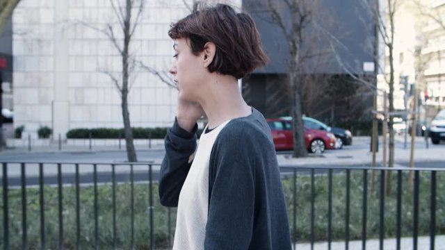 Side view of nervous woman walking and talking on smartphone. Stressed young lady strolling on street and discussing emergency questions via phone. Communication, technology, stress concept
