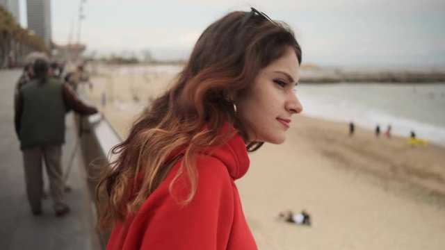 Close up shot of pretty hispanic girl in red hoodie dreamily resting on seafront 