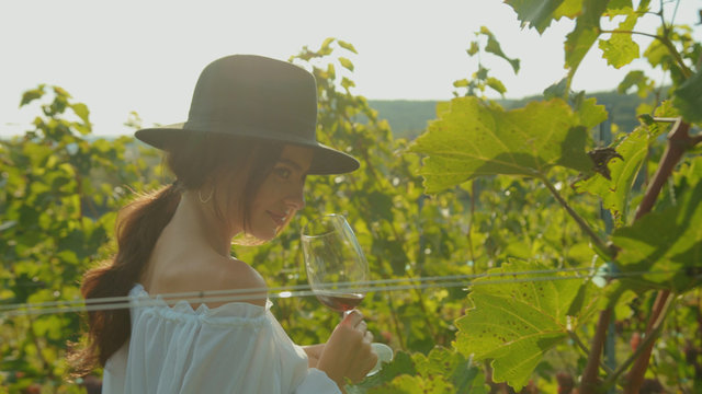Charming elegant young woman with glass of wine smiling running in grape vine alley enjoying summer vacation in vineyard.