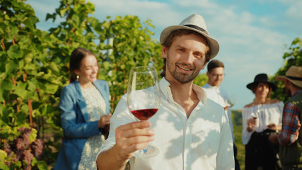 Happy young man with charming smile holding glass of pinot enjoying wine degustation experience with group of fashion friends. Tourism in vineyard.