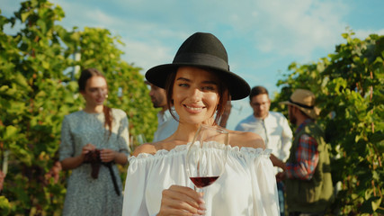 Fototapeta premium Spectacular young smiling woman in fashion outfit holding wineglass satisfied with beautiful nature and summer holiday event on vineyard.
