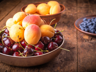 Ripe apricots, cherries and blueberries in bowls on a wooden table