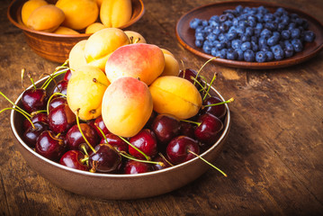 Ripe apricots, cherries and blueberries in bowls on a wooden table
