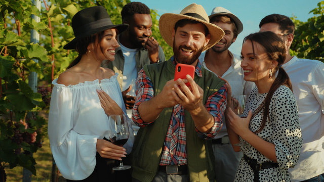 Happy group of friends watching fun content on smartphone display chilling out with wine drinks at vineyard on sunny day.