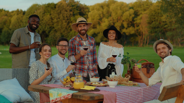 Beautiful happy friends toasting with red wine smiling enjoying summer celebratory picnic in the vineyard. Social gathering.