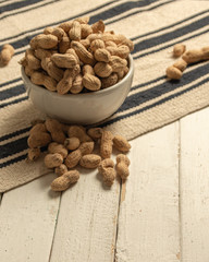 inshell peanuts in a white ceramic bowl on a cloth tablecloth and white wooden table