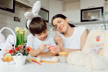 Mother and her son painting colorful easter eggs. Family celebrating Easter. Parent and kid play indoors. Decorated home and spring flowers.