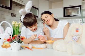 Obraz na płótnie Canvas Mother and her son painting colorful easter eggs. Family celebrating Easter. Parent and kid play indoors. Decorated home and spring flowers.