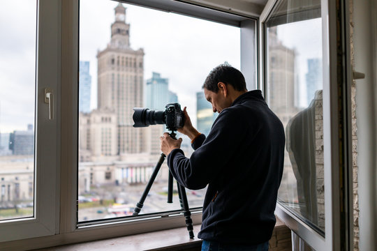 Young man in flat apartment with view of Warsaw, Poland cityscape taking picture with camera and tripod of famous Palace of Science and Culture building