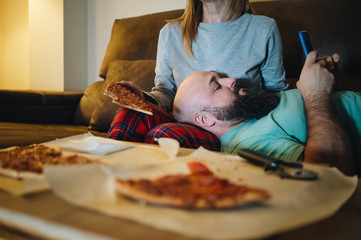 A couple of men and women eat pizza on the sofa in their living room at night while watching a movie on television. Concept of life as a couple at home.