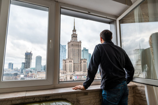 Young man standing looking in flat apartment with view of Warsaw, Poland famous Palace of Science and Culture cityscape building through window