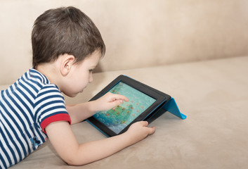 Toddler boy lie on the sofa with a tablet studying English. Distance learning during quarantine due to coronavirus pandemic concept. Stay home concept. Biohazard, 2019-nCoV, COVID-19, social distance.