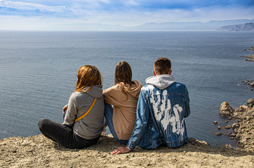 Fototapeta premium A guy and two girls are sitting on a mountain and looking at the sea. Travel and adventure, tourism.