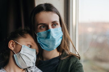 Tired mother and little daughter in medical sterile face mask sitting on sill looking at window,...