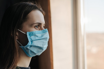 Side view young woman in wearing protective face mask sitting on window sill at home, looking...