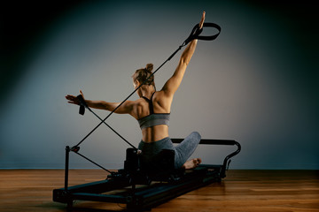Fototapeta na wymiar Young girl doing pilates exercises with a reformer bed. Beautiful slim fitness trainer on reformer gray background, low key, art light. Fitness concept