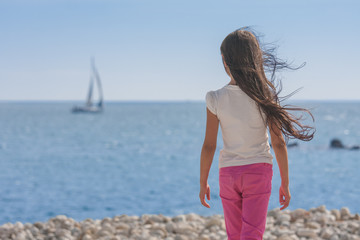 Back view of young brunette girl standing and looking at blue sea with her hair blowing.