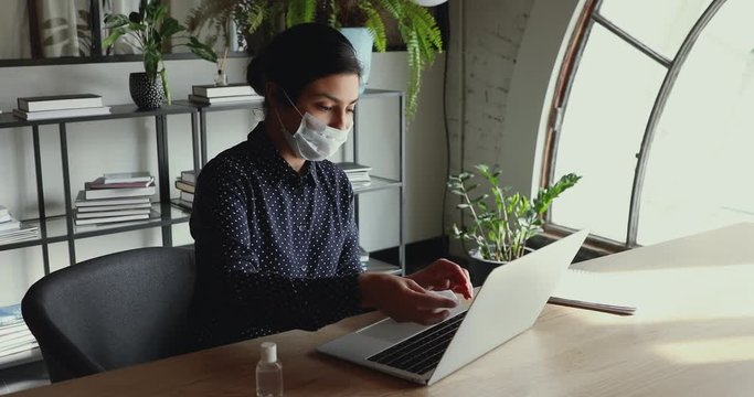 Indian business woman office worker wears medical face mask disinfecting laptop keyboard with sanitizer gel to stop coronavirus spreading. Sanitizing workplace devices for covid 19 prevention concept.