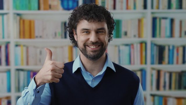 Happy middle age business entrepreneur smile cheerful pointing thumbs up in approval standing near bookshelves in a library. Positive gesturing.