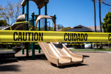 Yellow caution tape in english and spanish barring access to playgound equipment at a park. The access is denied due to the Coronavirus Pandemic
