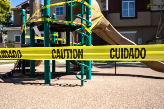 Yellow Caution Tape In English And Spanish Barring Access To Playgound Equipment At A Park. The Access Is Denied Due To The Coronavirus Pandemic