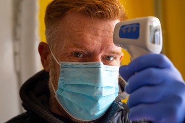 Man in a face mask and gloves checking body temperature with infrared forehead thermometer.