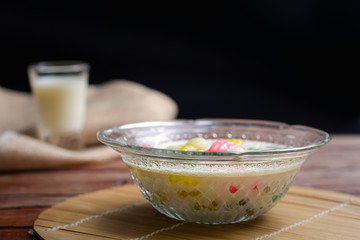 Colorful sweet rice gnocchi in fresh coconut milk in glass bowl on brown tablecloth on blackground. Delicious Thai desserts.