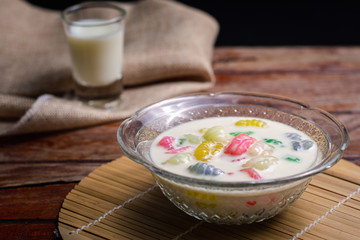 Colorful sweet rice gnocchi in fresh coconut milk in glass bowl on brown tablecloth on blackground. Delicious Thai desserts.