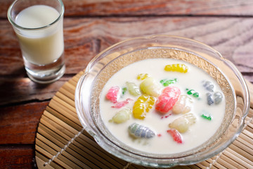 Colorful sweet rice gnocchi in fresh coconut milk in glass bowl on brown tablecloth on blackground. Delicious Thai desserts.