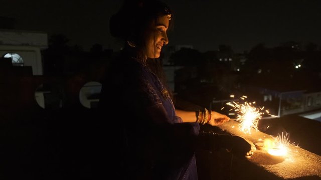 Young and beautiful Indian Bengali woman in Indian traditional dress is celebrating Diwali with fire crackers on a rooftop in darkness. Indian lifestyle and Diwali celebration