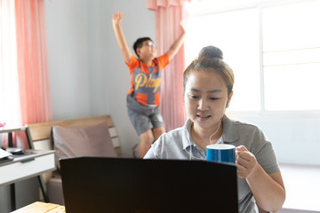 working from home. Asian mother using laptop and drink coffee and son make noise, Jump on the bed...