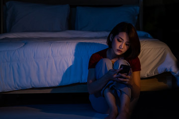 Depressed young beautiful Asian woman sitting alone on bedroom floor with looking at smartphone at...