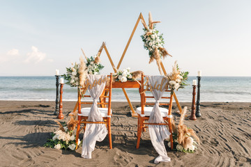 Triangular wedding arch in boho style in the coast of sea. Two chairs assigned to the bride and groom at a wedding setting - Powered by Adobe