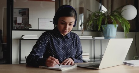 Female indian distant worker wears headphones video calling makes notes, elearning with online teacher, working from home office attending remote conference meeting. Videoconference training concept. - Powered by Adobe