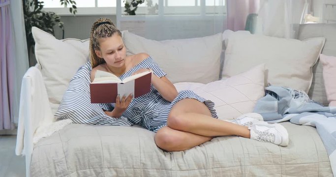 Young attractive teenage girl with breadlocks lying on sofa and reading book attentively. Positive sides of self-isolation at home during coronavirus epidemic