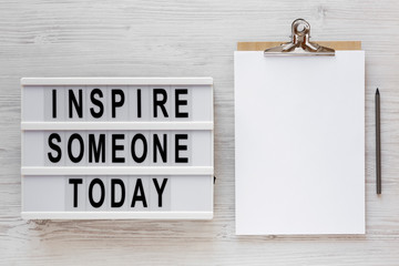 'Inspire someone today' words, clipboard with blank sheet of paper on a lightbox on a white wooden surface, top view. Overhead, from above, flat lay.